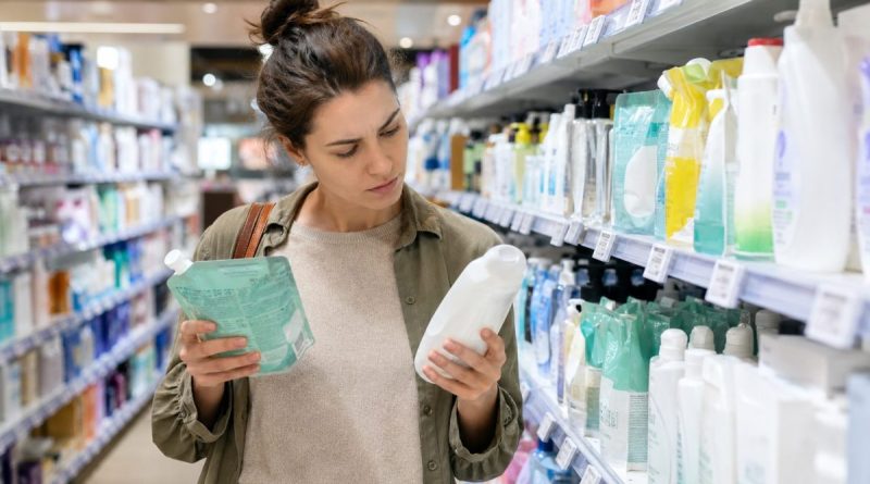 Consommatrice comparant attentivement plusieurs produits d’hygiène dans un rayon de supermarché, tenant une recharge souple et un flacon classique afin de vérifier les prix affichés.