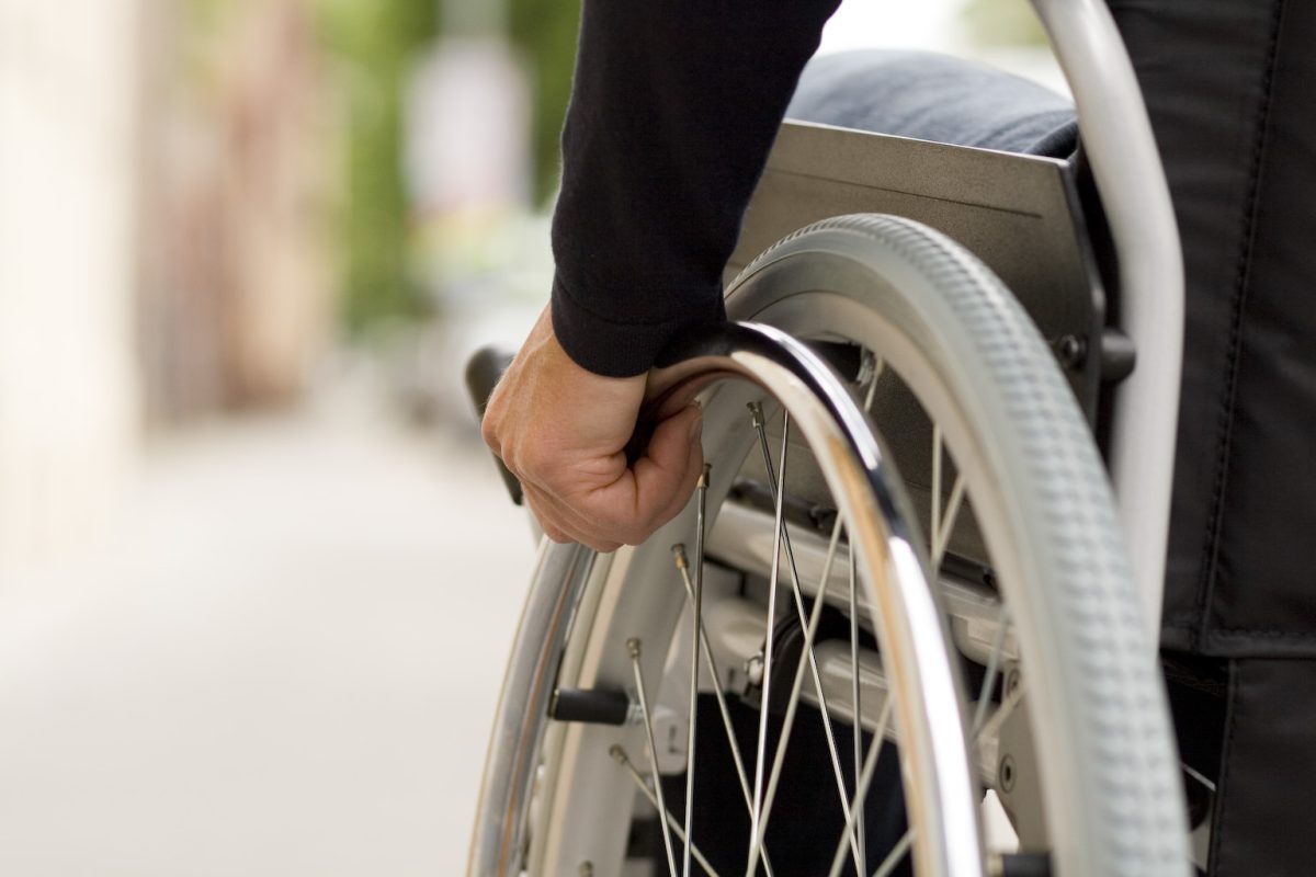 Personne en fauteuil roulant se déplaçant de manière autonome sur un chemin urbain, avec un gros plan sur la main actionnant la roue. Cette image illustre le quotidien des personnes à mobilité réduite, l’accessibilité, le handicap et les questions liées aux déplacements en fauteuil roulant dans l’espace public.