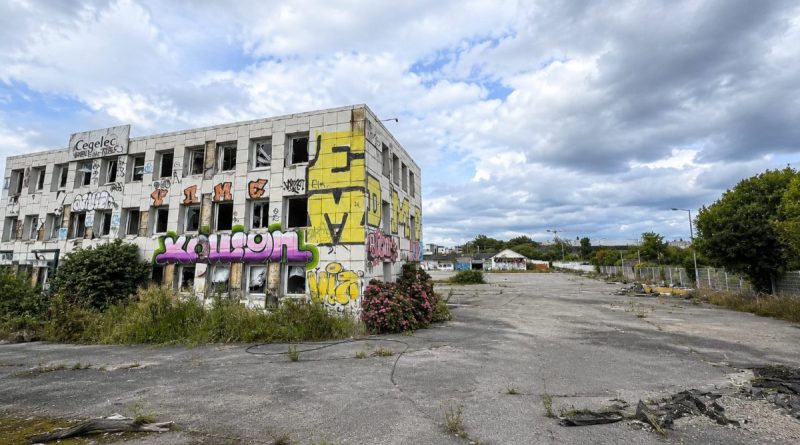 Ancien bâtiment industriel abandonné recouvert de graffitis colorés au bord d'une vaste esplanade asphaltée envahie par la végétation. Le site désaffecté présente des fenêtres brisées, des façades dégradées et un grand espace vide sous un ciel nuageux. Cette friche urbaine située au nord de Rennes est appelée à être transformée dans le cadre d'un important projet d'aménagement.