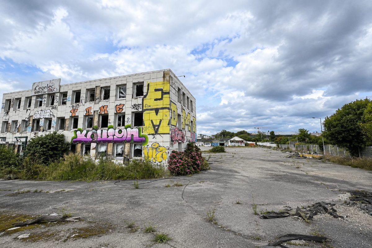 Ancien bâtiment industriel abandonné recouvert de graffitis colorés au bord d'une vaste esplanade asphaltée envahie par la végétation. Le site désaffecté présente des fenêtres brisées, des façades dégradées et un grand espace vide sous un ciel nuageux. Cette friche urbaine située au nord de Rennes est appelée à être transformée dans le cadre d'un important projet d'aménagement.