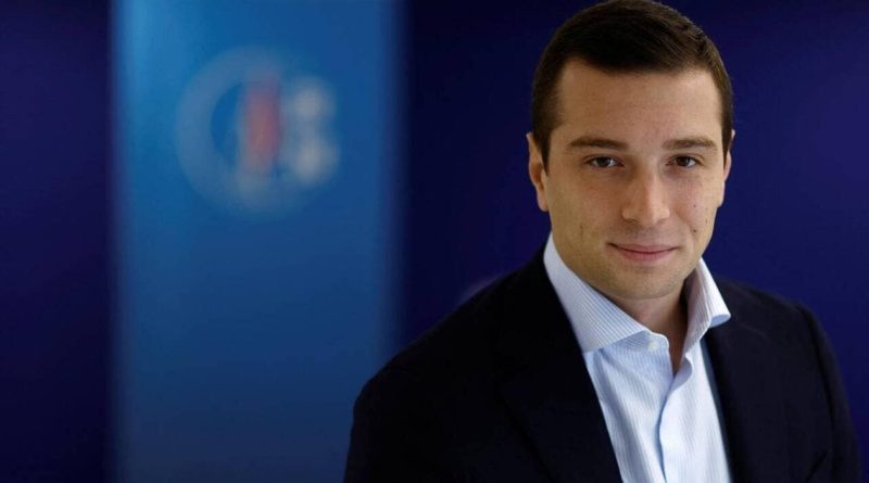 Portrait de Jordan Bardella en costume sombre et chemise claire, regardant l’objectif dans un environnement institutionnel aux tons bleus. L’image illustre une actualité politique liée au Rassemblement National, aux médias et au débat sur le pluralisme dans l’audiovisuel public.