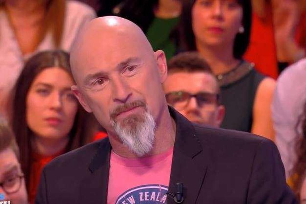 Homme chauve à la barbe blanche participant à une émission de télévision en public, vêtu d'une veste sombre et d'un tee-shirt coloré, observant attentivement une discussion sur un plateau entouré de spectateurs.