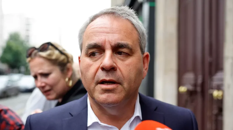 Responsable politique français en costume sombre répondant aux questions de journalistes à l'extérieur d'un bâtiment officiel. Entouré de micros et sous l'attention des médias, il apparaît dans un contexte de débat public et de controverse politique nationale, tandis que plusieurs personnes sont visibles en arrière-plan dans une rue urbaine.