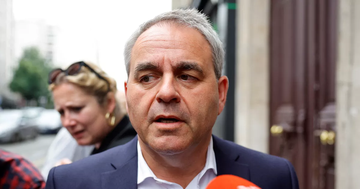Responsable politique français en costume sombre répondant aux questions de journalistes à l'extérieur d'un bâtiment officiel. Entouré de micros et sous l'attention des médias, il apparaît dans un contexte de débat public et de controverse politique nationale, tandis que plusieurs personnes sont visibles en arrière-plan dans une rue urbaine.