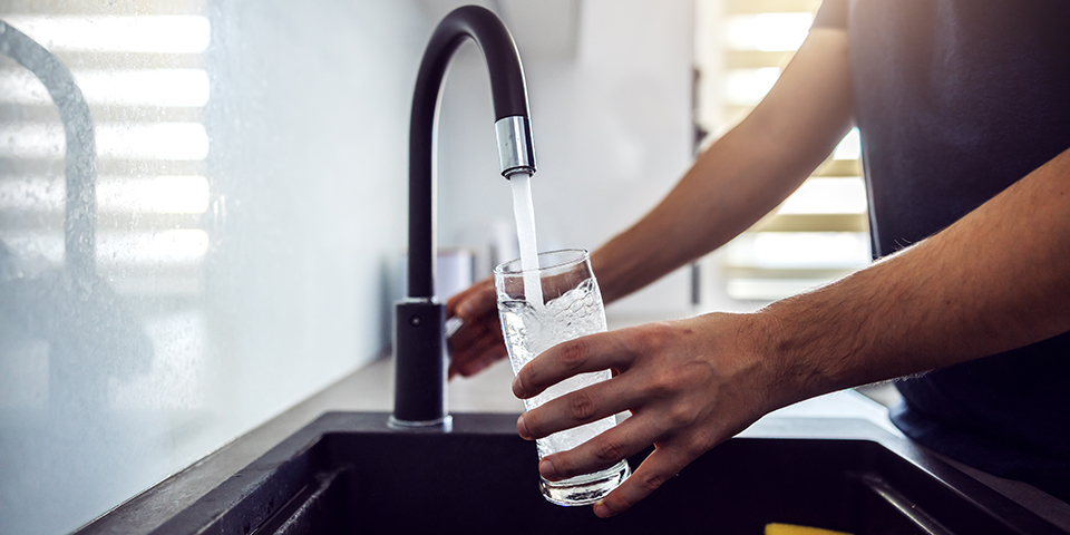 Une personne remplit un verre d'eau au robinet dans une cuisine moderne pendant un épisode de canicule. Lorsque les fortes chaleurs réchauffent les canalisations, l'eau potable peut dépasser 25 °C et favoriser le développement de micro-organismes. Cette image illustre les précautions à prendre avant de boire l'eau du robinet en période de fortes températures.