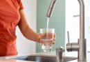 Une personne remplit un verre d'eau au robinet dans une cuisine pendant une période de canicule. L'eau potable peut dépasser 25 °C dans les canalisations lors de fortes chaleurs, favorisant le développement de micro-organismes. Illustration des risques liés à une eau du robinet trop chaude et des précautions recommandées pour préserver la qualité de l'eau potable.