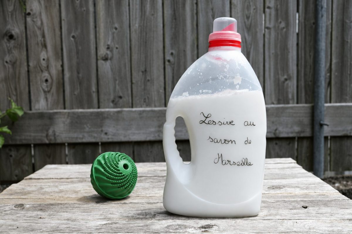 Bidon réutilisé contenant de la lessive maison au savon de Marseille, posé sur une table en bois à côté d’une boule de lavage verte.