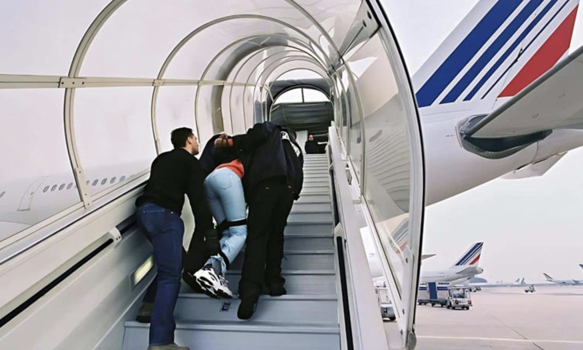 Un ressortissant algérien escorté par des agents des forces de l'ordre monte de force dans un avion dans le cadre d'une procédure d'expulsion liée à une OQTF après plusieurs refus d'embarquer vers l'Algérie.