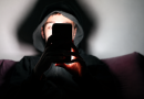Jeune homme portant une capuche noire, plongé dans l’obscurité et regardant un smartphone dont la lumière éclaire son visage et ses mains, illustration évoquant l’isolement, la détresse psychologique et les échanges avec une intelligence artificielle comme ChatGPT.