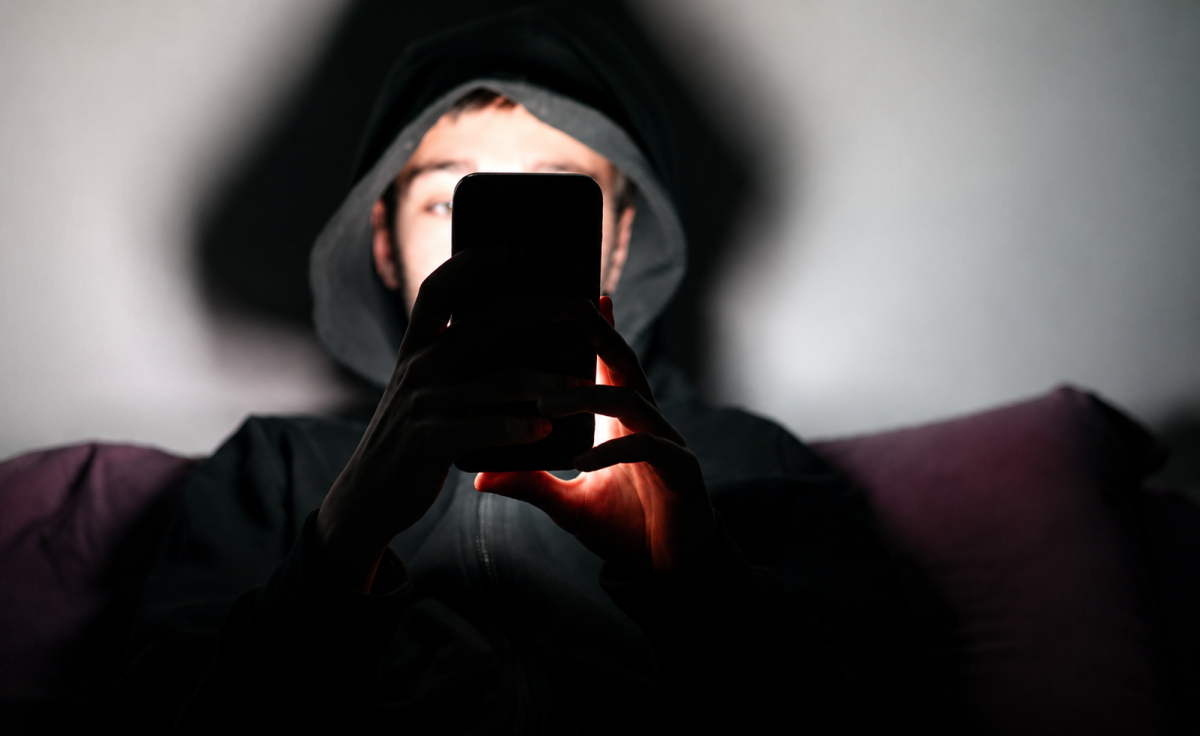Jeune homme portant une capuche noire, plongé dans l’obscurité et regardant un smartphone dont la lumière éclaire son visage et ses mains, illustration évoquant l’isolement, la détresse psychologique et les échanges avec une intelligence artificielle comme ChatGPT.