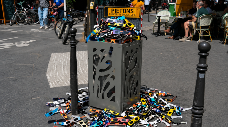 « Une poubelle publique déborde de lunettes de protection utilisées pour observer l’éclipse solaire, tandis que de nombreuses autres lunettes sont abandonnées sur le trottoir, illustrant la question des déchets, du gaspillage et de l’impact environnemental des objets utilisés pendant une très courte durée. »