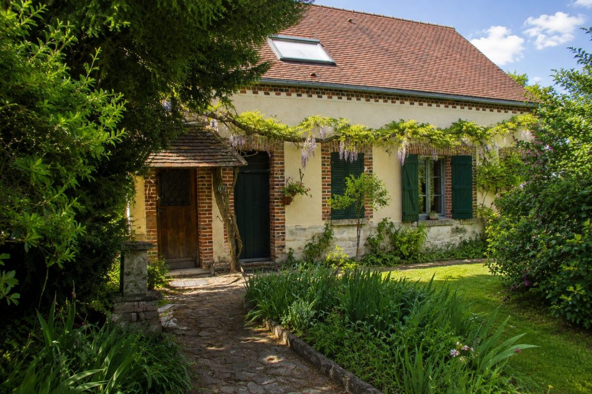 Illustration photoréaliste d'une maison de campagne inspirée de la résidence secondaire de Jean-Luc Mélenchon, située près de Montargis dans le Loiret, entourée d'un jardin arboré et d'une végétation luxuriante.