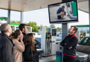 Illustration photoréaliste d'une station-service moderne avec plusieurs pompes à carburant. Des clients, visiblement choqués, regardent un écran diffusant une image volontairement floutée d'une scène d'incivilité, tandis qu'un employé observe la situation les bras croisés. Cette illustration accompagne un article consacré à la décision d'un Super U d'afficher une capture de vidéosurveillance pour dénoncer le comportement inapproprié d'un client.
