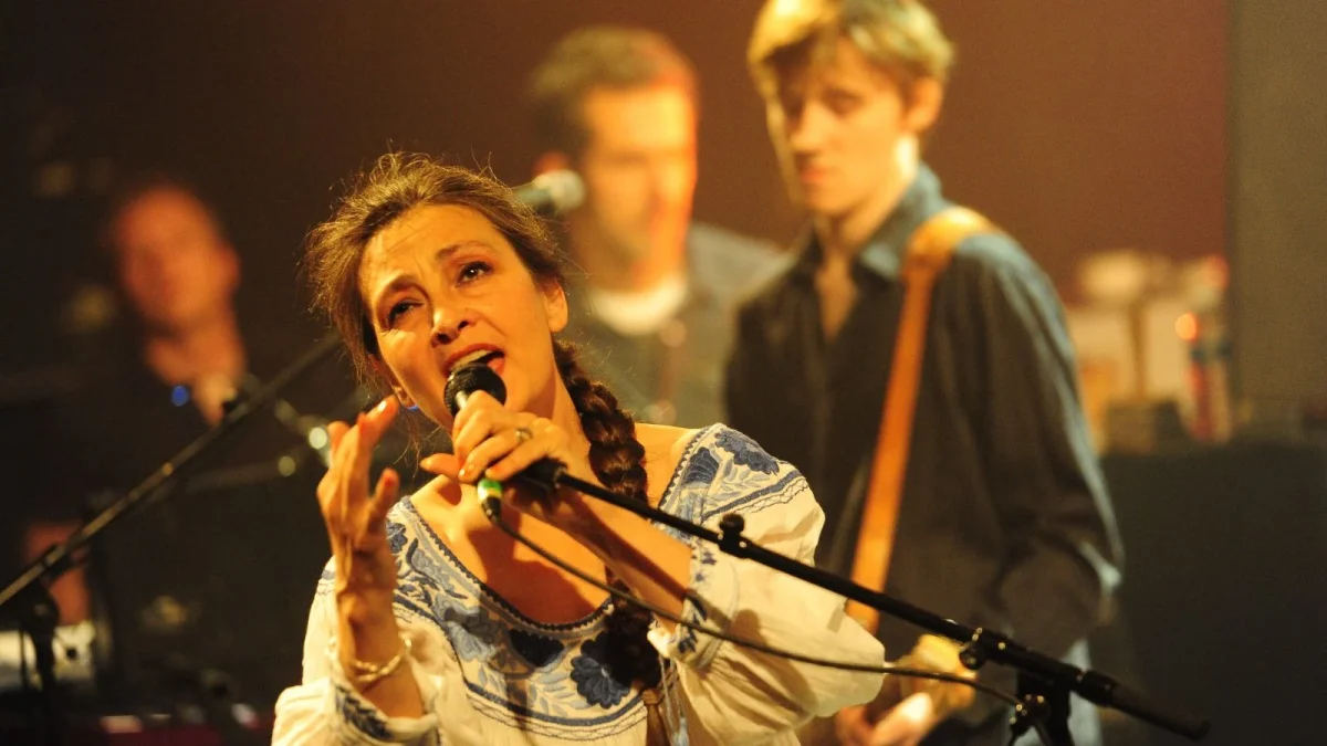 Catherine Ringer en concert sur scène, chantant au micro devant ses musiciens, lors d’une performance illustrant sa carrière avec les Rita Mitsouko et en solo.
