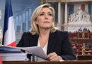 Marine Le Pen devant des documents budgétaires, avec l’Assemblée nationale et le drapeau français en arrière-plan, illustration de sa stratégie concernant le budget 2027 avant l’élection présidentielle.