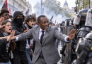 Illustration de Bally Bagayoko, maire de Saint-Denis, lors de la manifestation lycéenne du 6 octobre 2026 à Paris, bras tendus face aux policiers en tenue antiémeute, au milieu de jeunes manifestants, dans un climat de fortes tensions et de fumée.