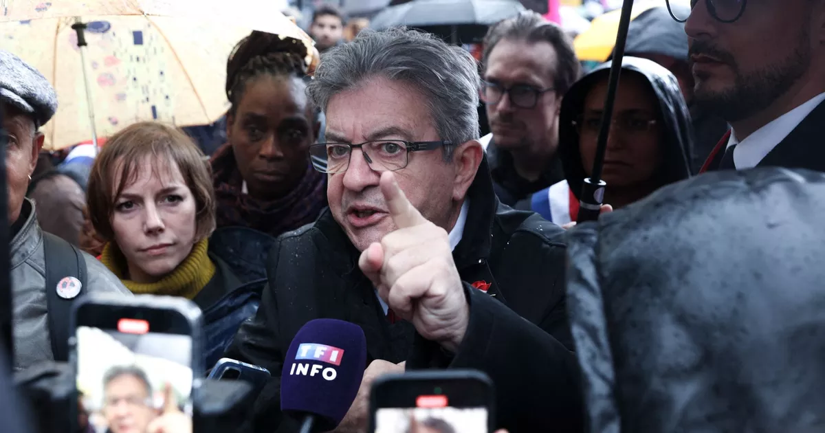Jean-Luc Mélenchon au milieu d’une foule sous la pluie, entouré de militants, de journalistes et de téléphones portables, s’exprimant avec vigueur en pointant du doigt devant un micro de TF1 Info lors d’un rassemblement politique.