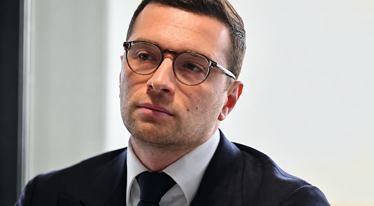 Portrait de Jordan Bardella portant des lunettes et un costume sombre, dans le cadre de l’affaire l’opposant à Mediapart en 2026.