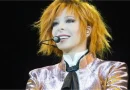 Mylène Farmer sur scène en concert, avec ses cheveux roux, un micro-casque et une veste brillante aux tons rose et doré.