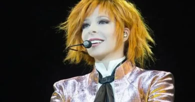Mylène Farmer sur scène en concert, avec ses cheveux roux, un micro-casque et une veste brillante aux tons rose et doré.
