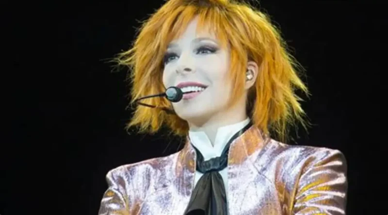 Mylène Farmer sur scène en concert, avec ses cheveux roux, un micro-casque et une veste brillante aux tons rose et doré.