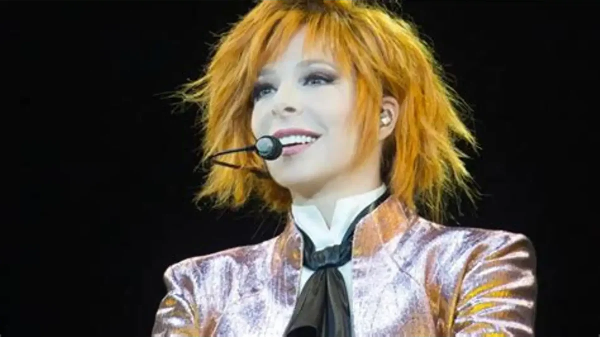 Mylène Farmer sur scène en concert, avec ses cheveux roux, un micro-casque et une veste brillante aux tons rose et doré.