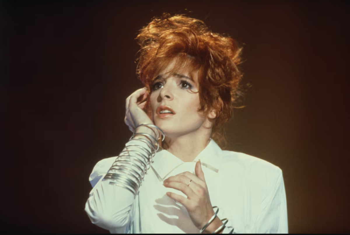 Mylène Farmer jeune sur scène, cheveux roux relevés et vêtue de blanc, pendant les premières années de sa carrière.