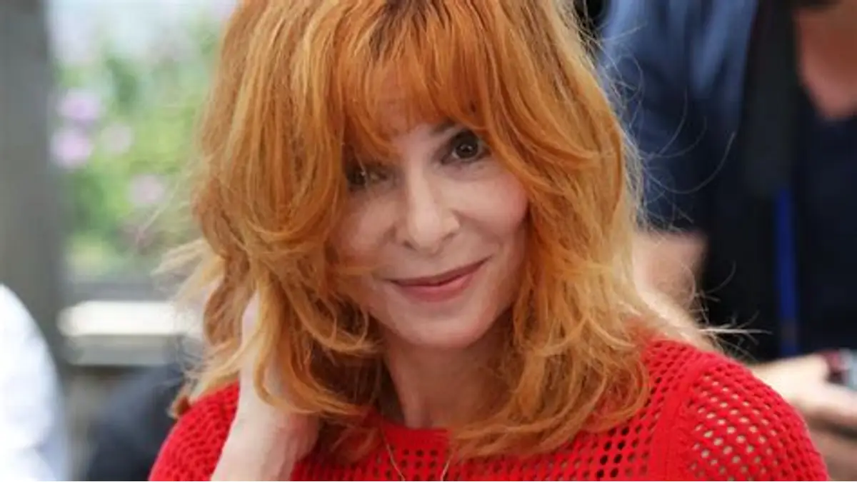 Mylène Farmer souriante, les cheveux roux, portant un haut rouge lors d’une apparition publique.